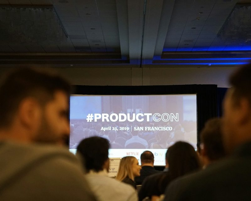 Conference audience facing a large projection screen in a darkened hall